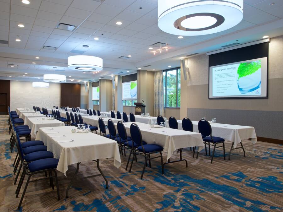 A conference room with long tables, chairs, and a large screen displaying a presentation.
