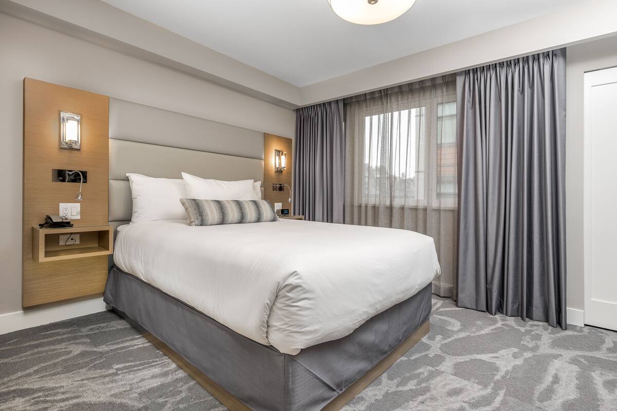 Een moderne hotelkamer met een groot bed, een nachtkastje met lamp, een wandlamp en uitzicht op de stad door dunne gordijnen.