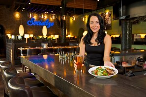 A bar area with a neon sign, bar stools, and a person holding a drink. - Coast Tsawwassen Inn (Delta)