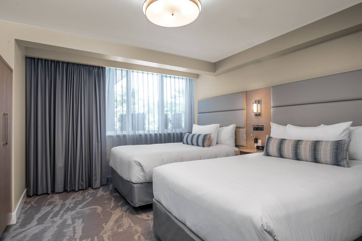 Hotelkamer met twee bedden, een groot raam met gordijnen, een hoofdeinde met verlichting en een vloerbedekking.