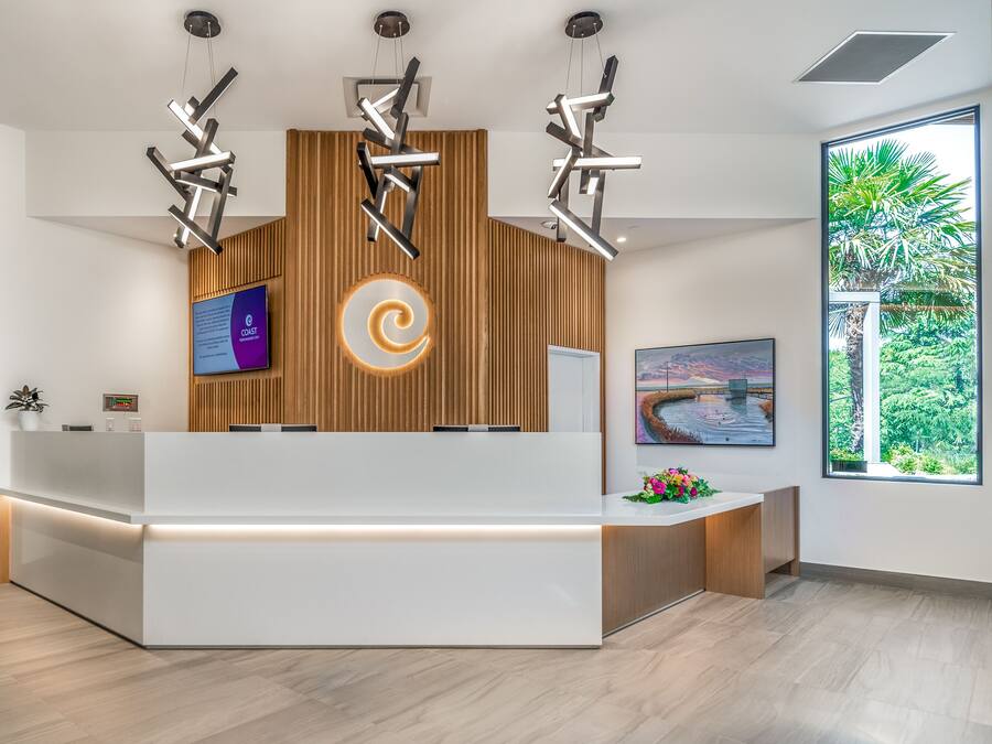 A modern reception desk with a wooden panel backdrop, a digital screen, and a large window offering a view of greenery.