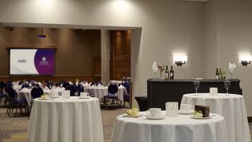 A conference room with round tables, chairs, a projector screen, and a presentation area.