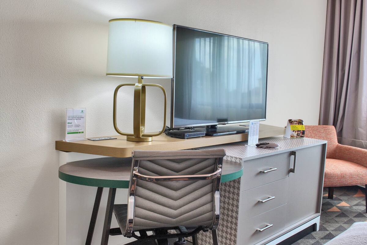 A hotel room with a desk, a television, a lamp, a chair, and a sofa.