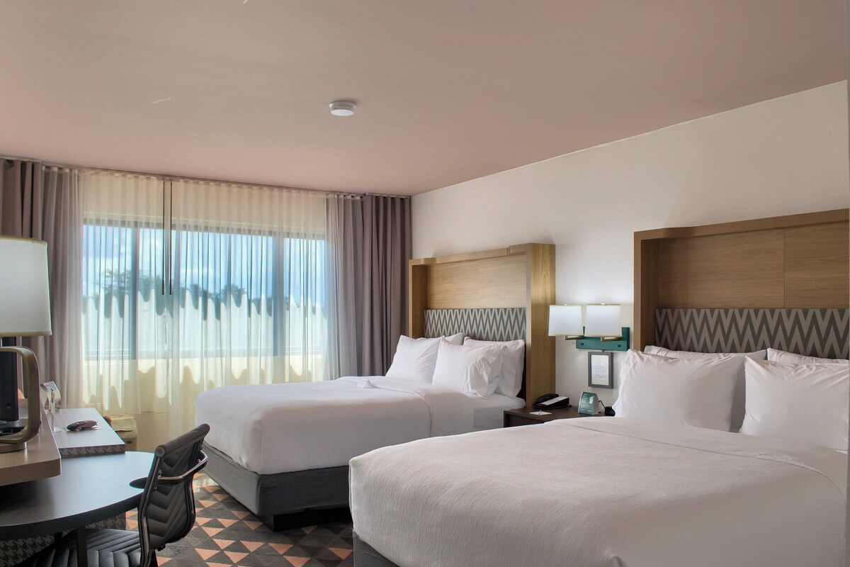 A hotel room with two beds, a desk with a chair, a telephone, and a window with curtains.