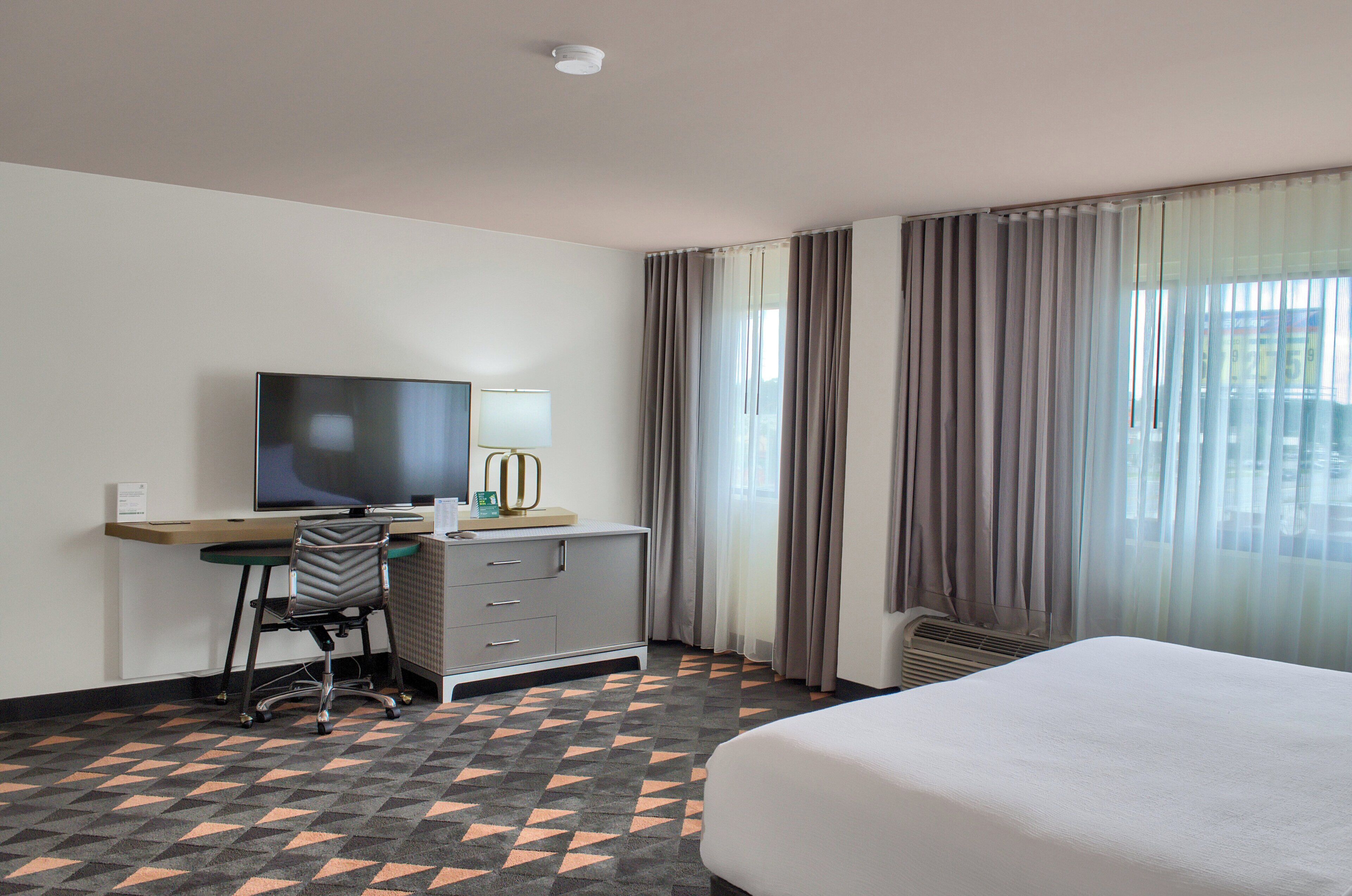 Habitación de hotel con cama, escritorio con silla, televisor, cómoda y ventana con cortinas.