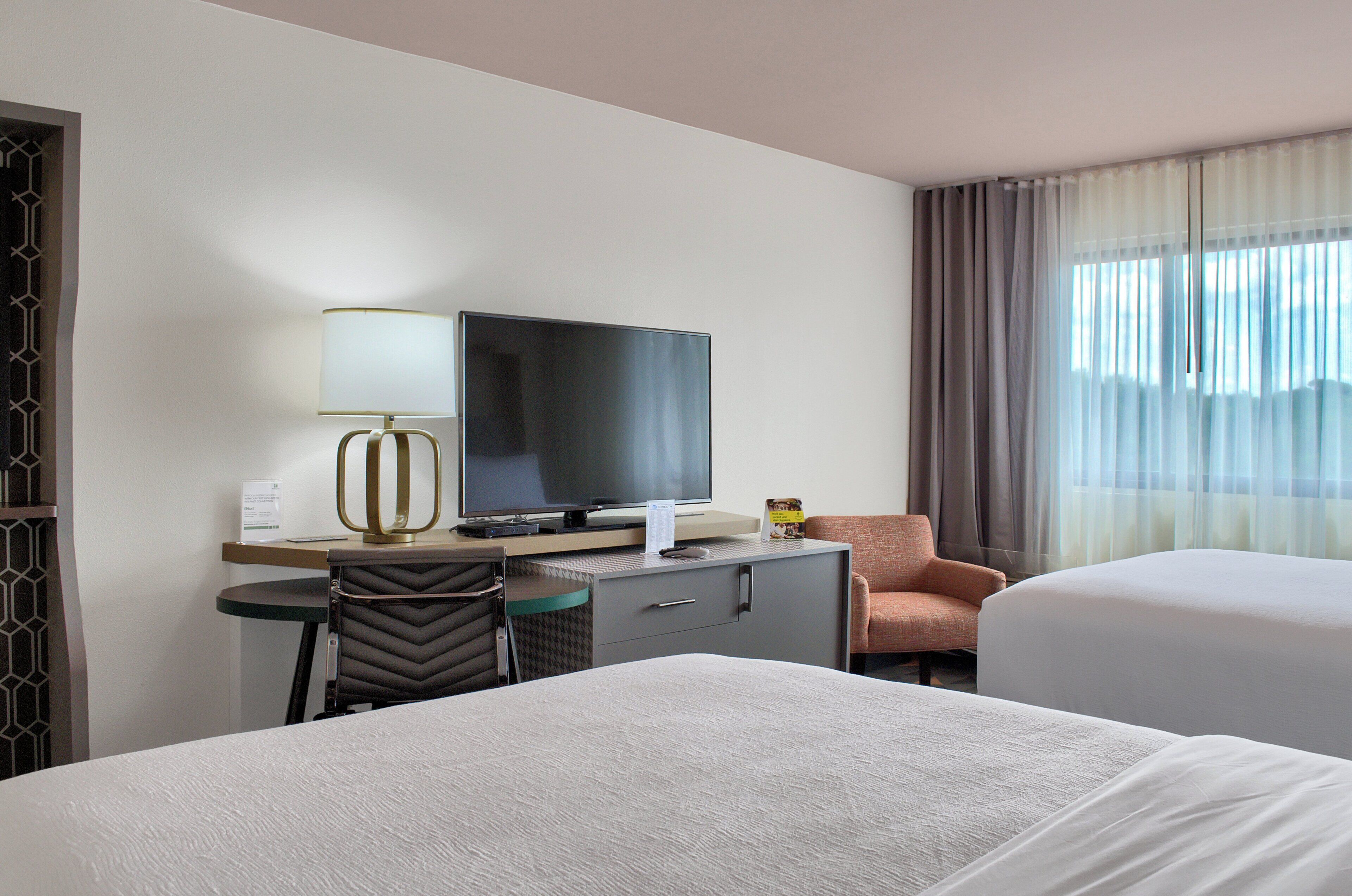 Una camera d'hotel con un letto, una scrivania con televisore, una sedia e una finestra con tende.