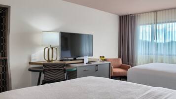 Una camera d'hotel con un letto, una scrivania con televisore, una sedia e una finestra con tende.