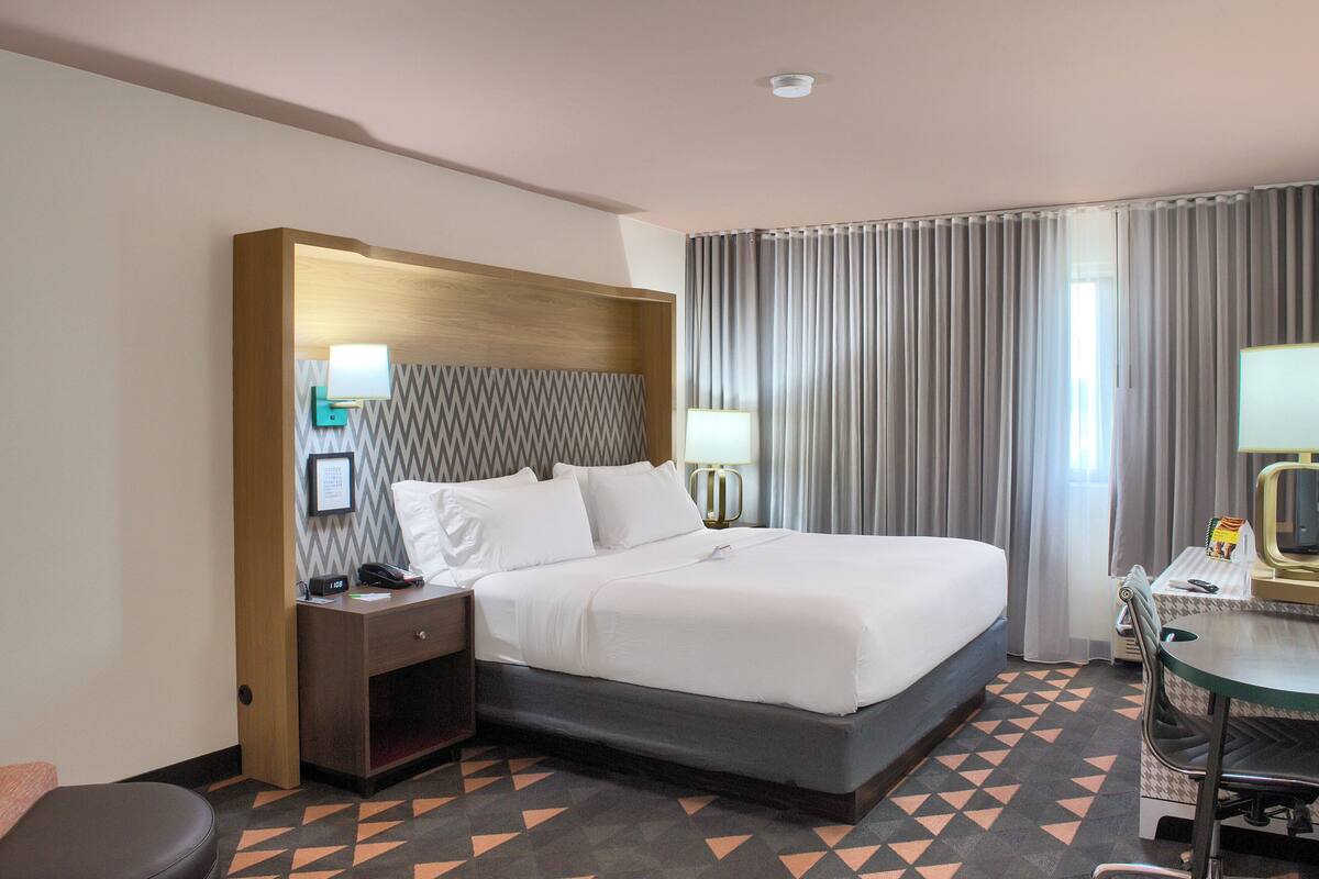A hotel room with a large bed, a desk with a chair, a small table, a lamp, and a window with curtains.