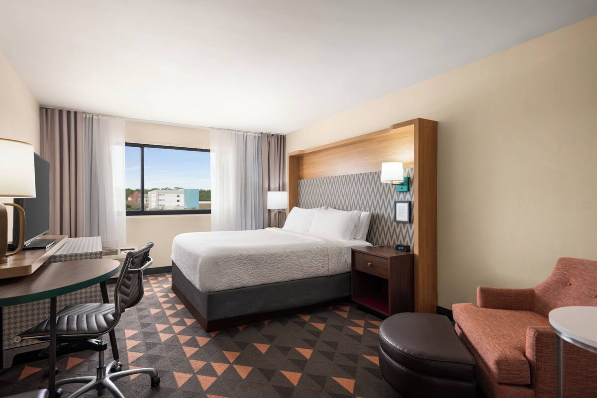 A hotel room with a bed, desk, chair, and a view of the city.