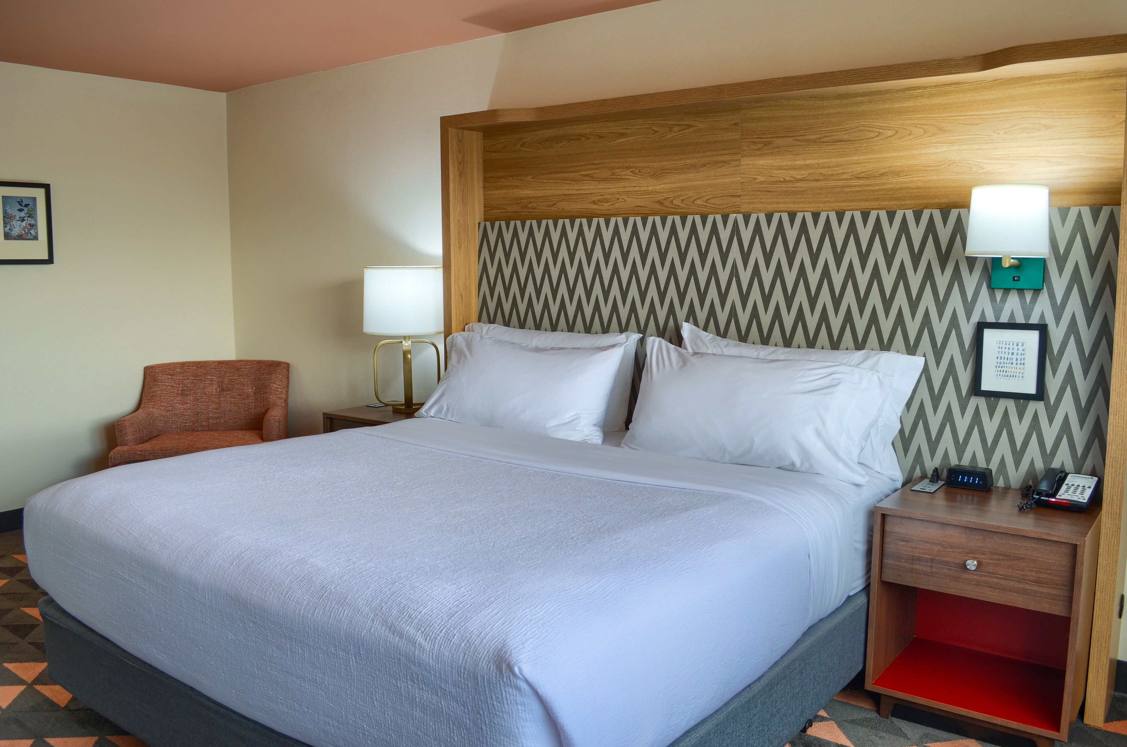 A hotel room with a large bed, a wooden headboard, a nightstand with a lamp, a chair, and a red shelf.