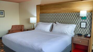A hotel room with a large bed, a wooden headboard, a nightstand with a lamp, a chair, and a red shelf.