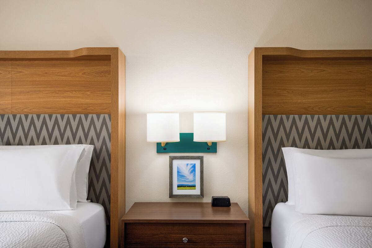 A hotel room with two beds, a nightstand with a framed picture, and two wall-mounted lamps.