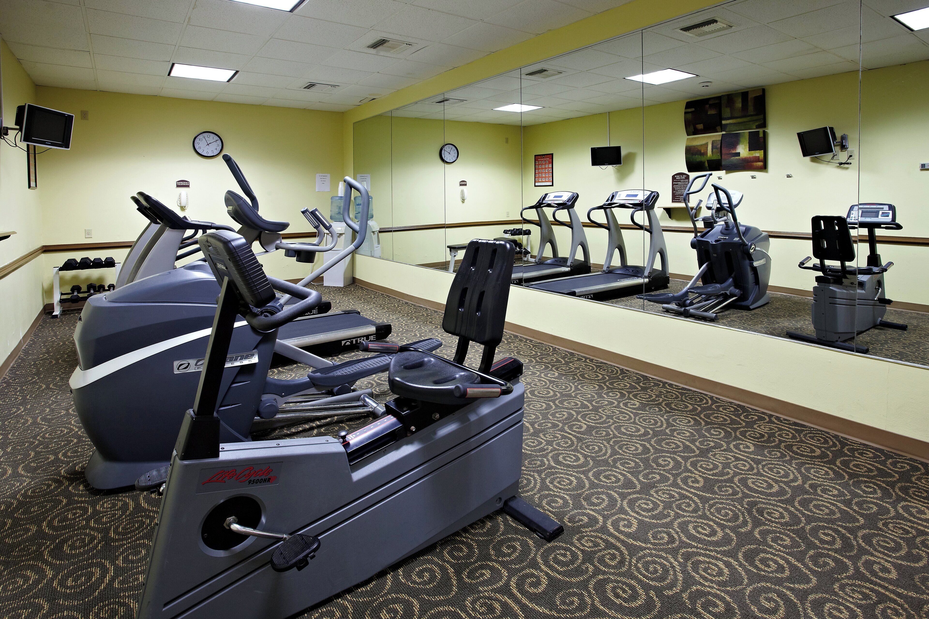 A gym with exercise equipment, including treadmills and an elliptical machine, carpeted flooring, and a mirror on the wall.