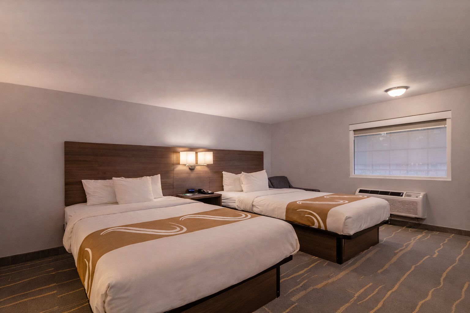 A hotel room with two beds, a nightstand, a desk, a chair, a window, and an air conditioning unit.