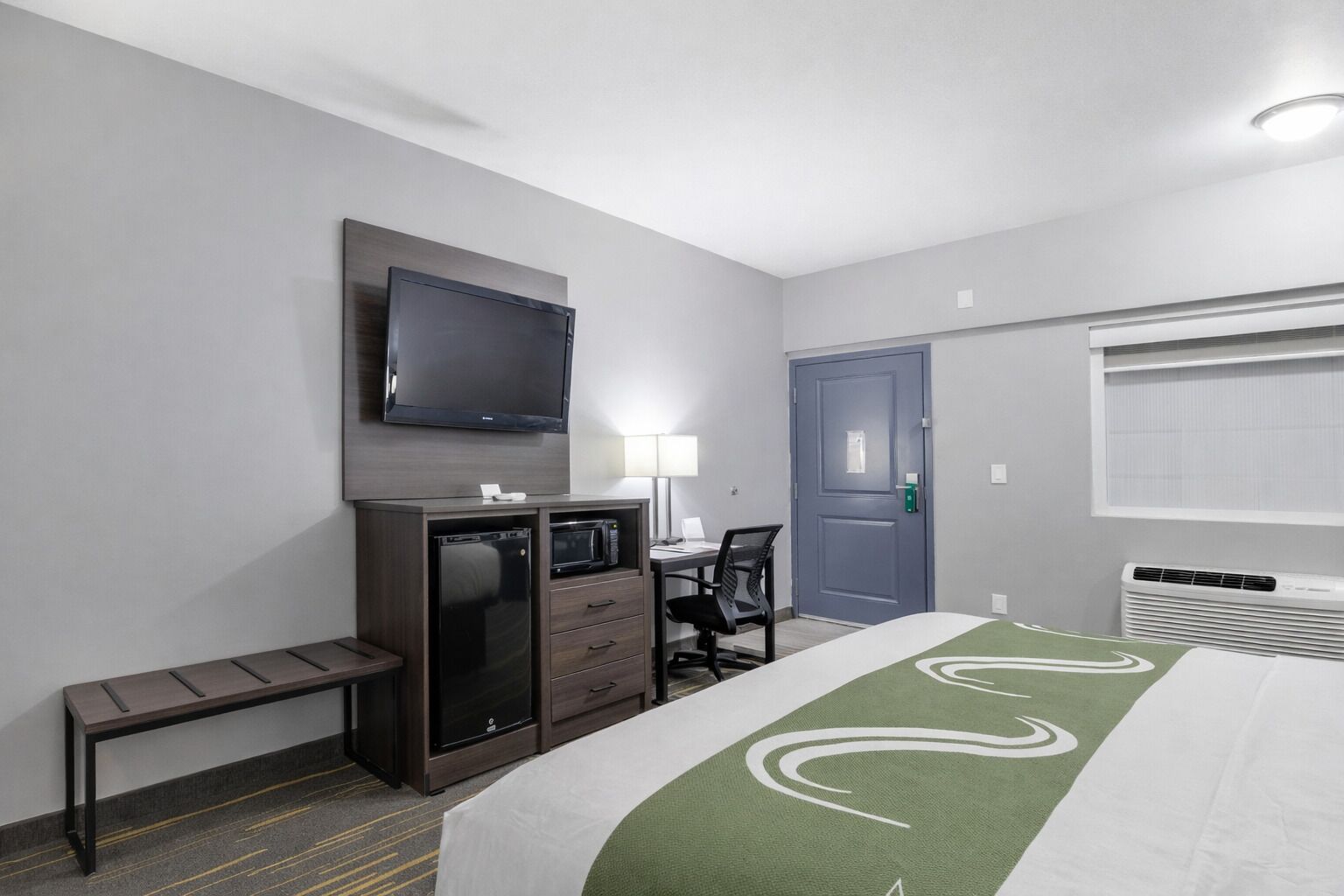Habitación de hotel con cama, escritorio y silla, televisor montado en la pared, microondas, refrigerador, banco, puerta y ventana con persianas.