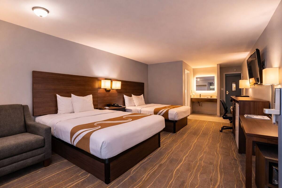 A hotel room with two beds, a desk, a chair, a TV, and a bathroom visible through an open door.