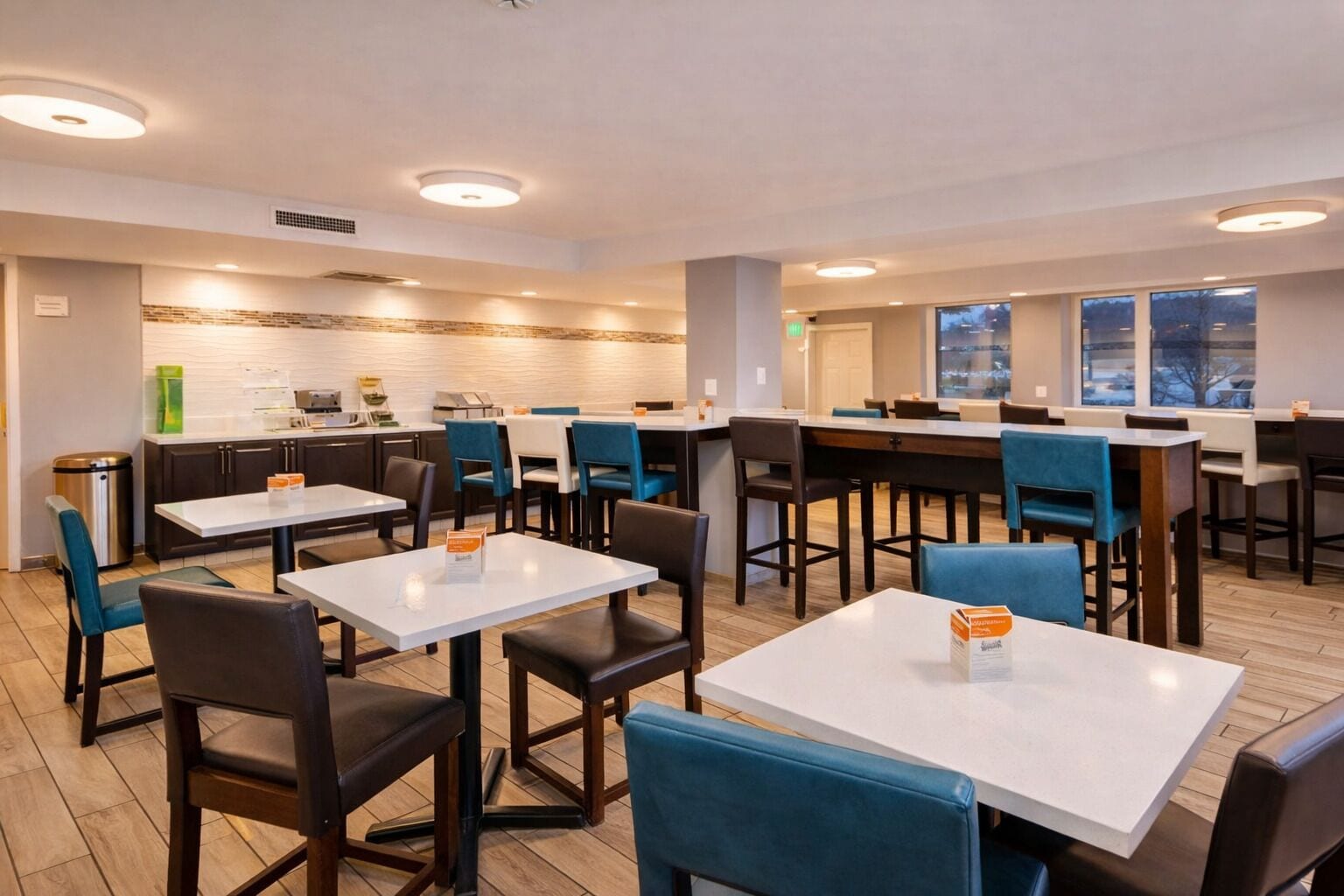 Un comedor moderno con mesas blancas y sillas azules, una cocina al fondo y ventanas amplias con vistas.
