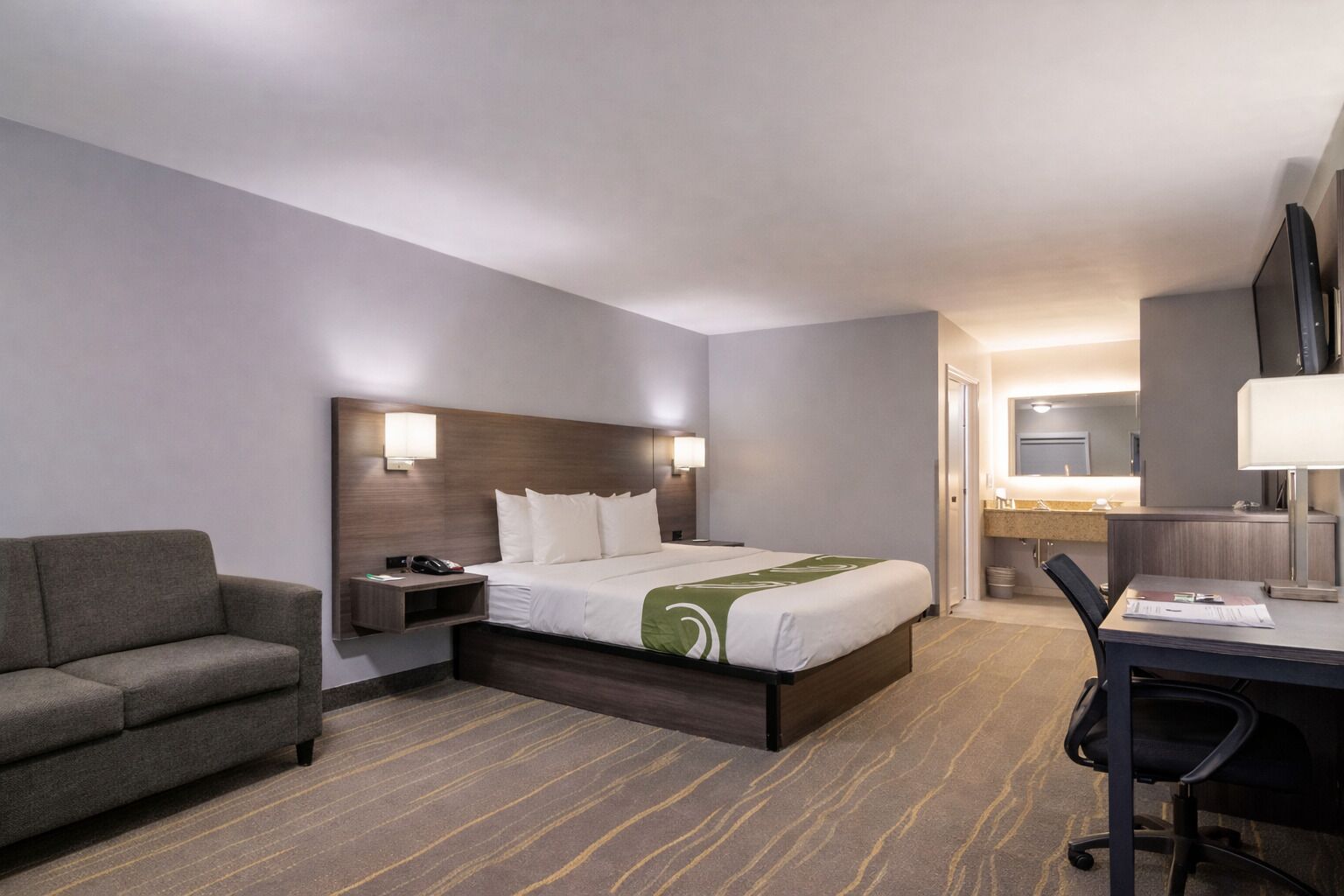 Habitación de hotel con cama, escritorio, silla, sofá, mesita de noche y baño visible a través de una puerta abierta.