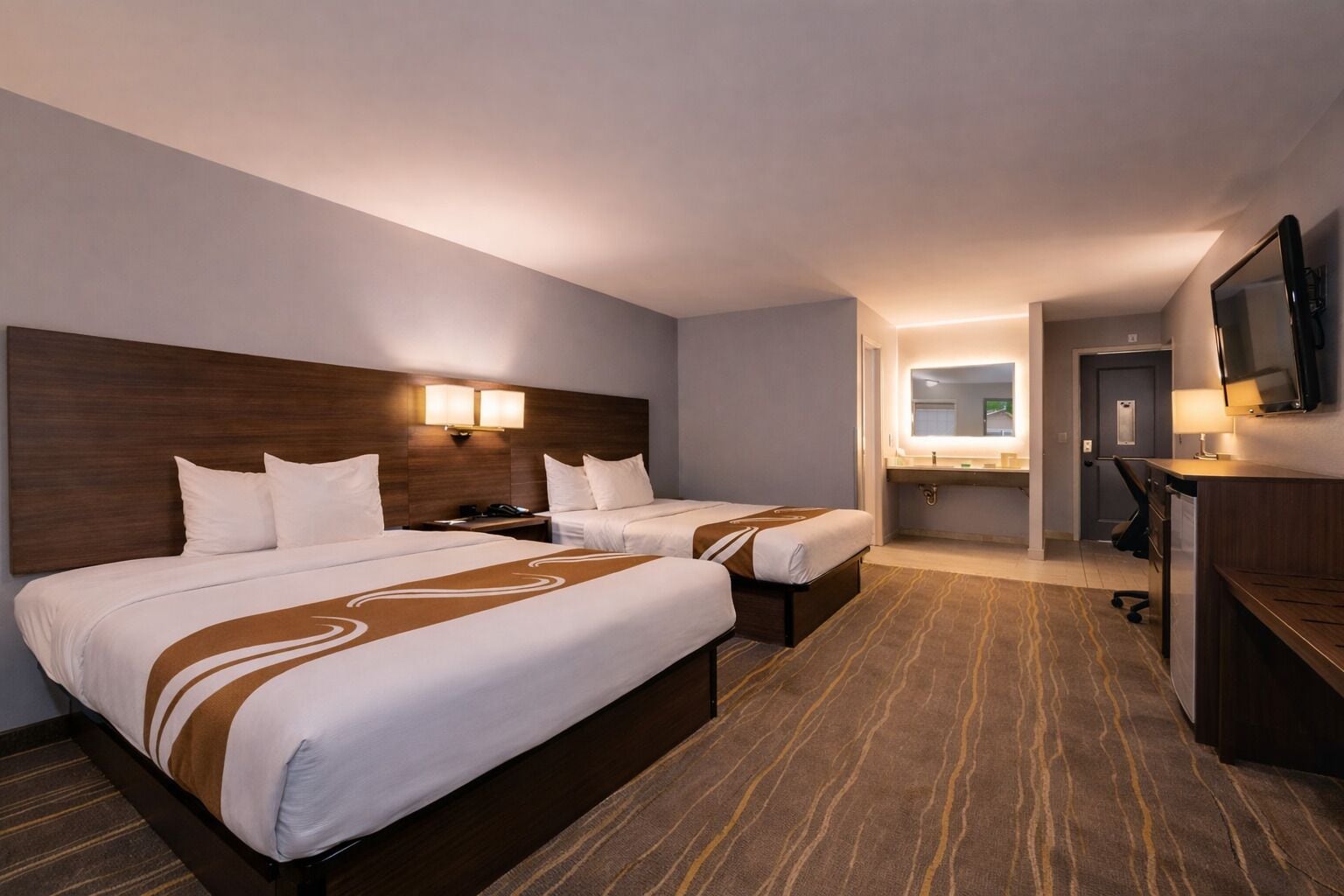 A hotel room with two beds, a desk, a TV, and a bathroom visible through an open door.