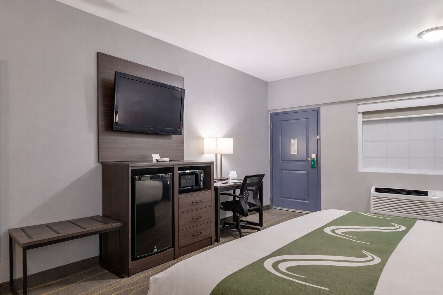 Quarto de hotel com cama, escrivaninha com cadeira, televisão na parede, porta e ar condicionado.