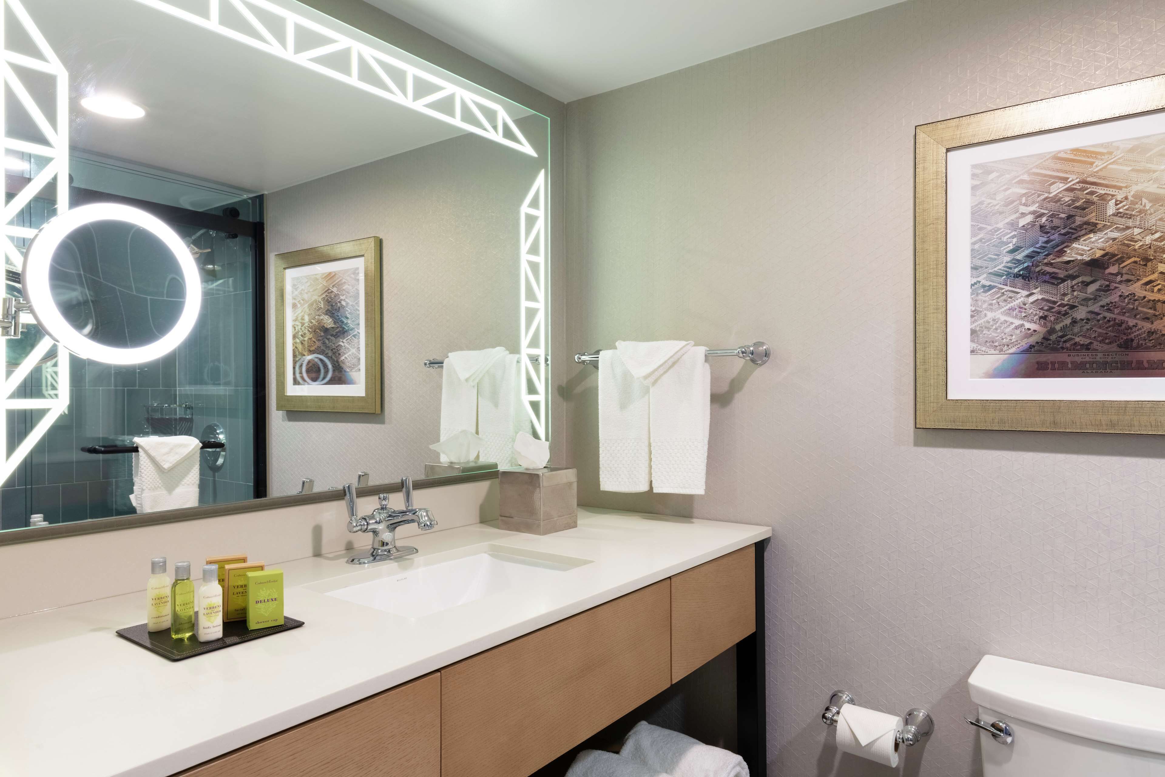 A modern bathroom with a large mirror, a sink with toiletries, a toilet, and a framed artwork on the wall.