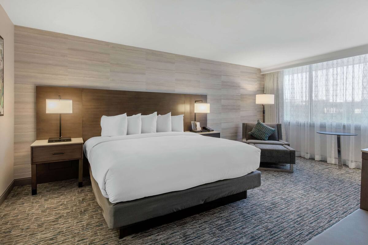 A modern hotel room with a large bed, two bedside tables with lamps, a sofa, and a view of the city through large windows.