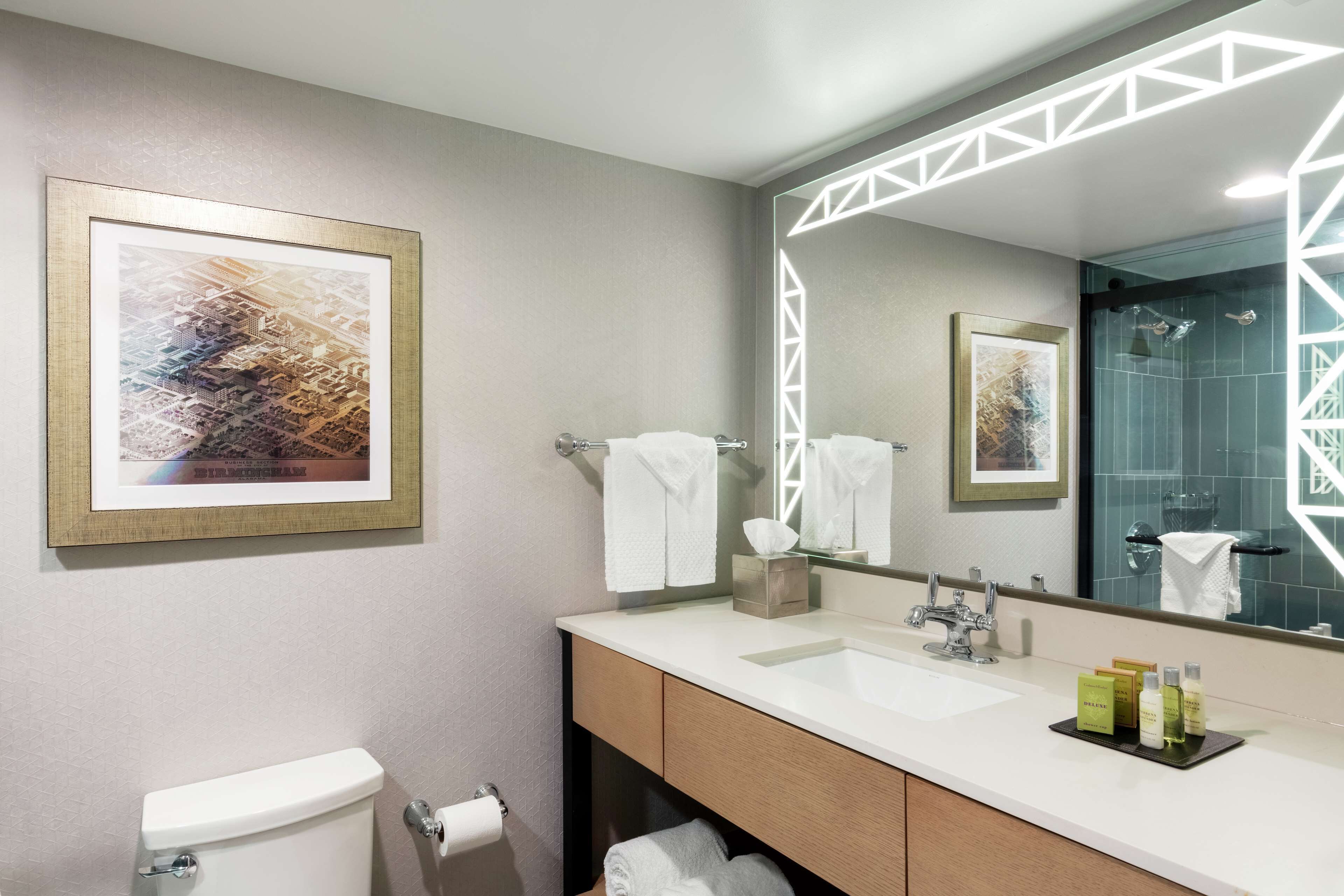 A modern bathroom with a toilet, sink, and large mirror. There is a framed abstract painting on the wall and a towel rack with towels.