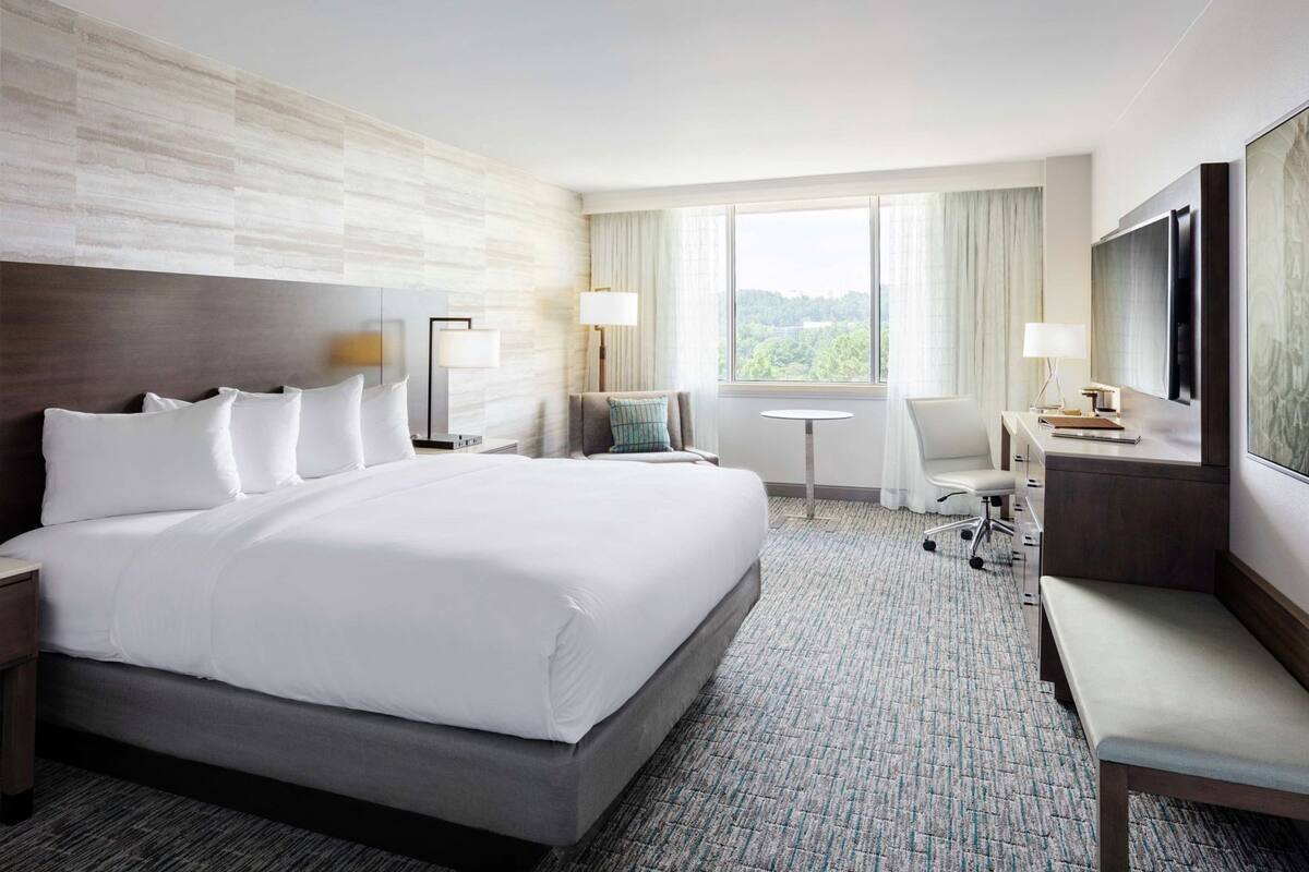 A hotel room with a large bed, bedside tables, a desk, a chair, a TV, and a window with a view.