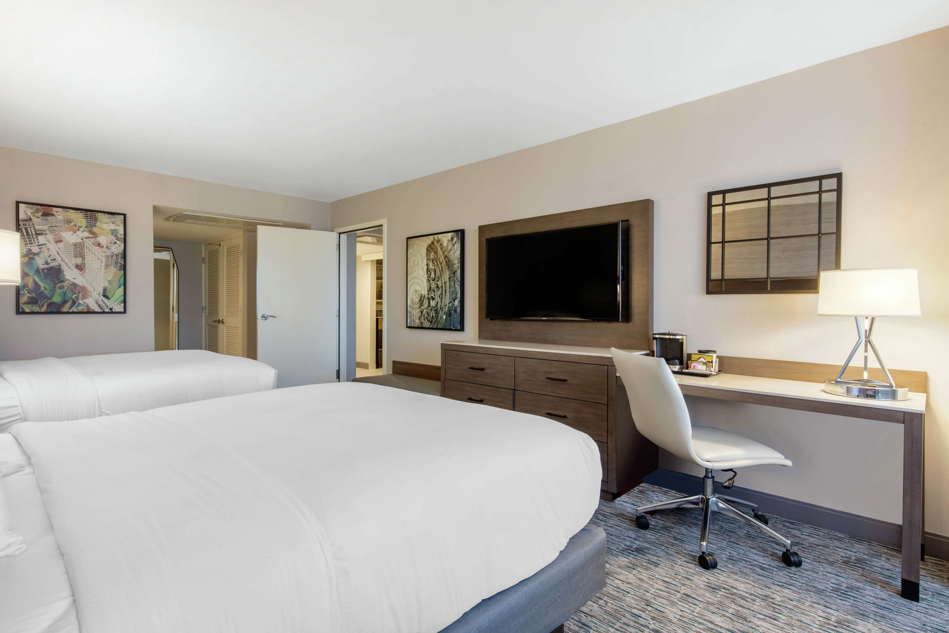 A hotel room with two beds, a desk with a chair, a flat-screen TV, and artwork on the walls.