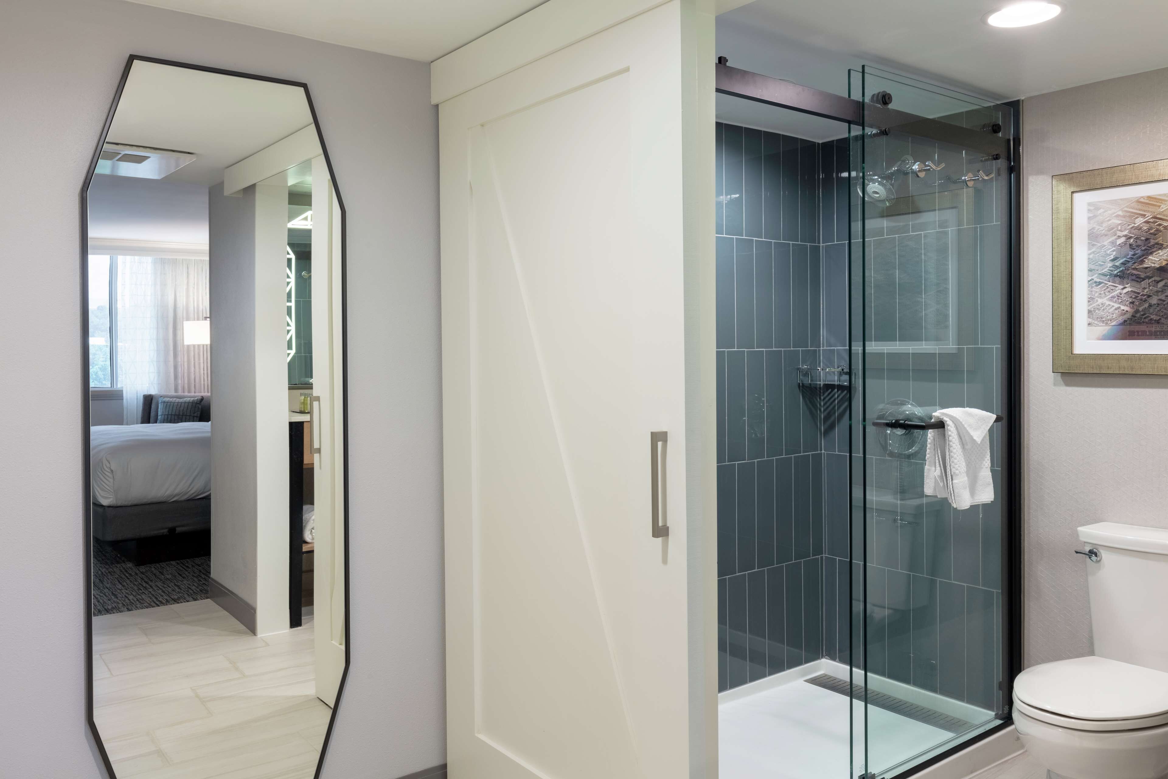 A modern bathroom with a glass shower enclosure, a white toilet, and a large mirror reflecting a bedroom with a bed and a window.