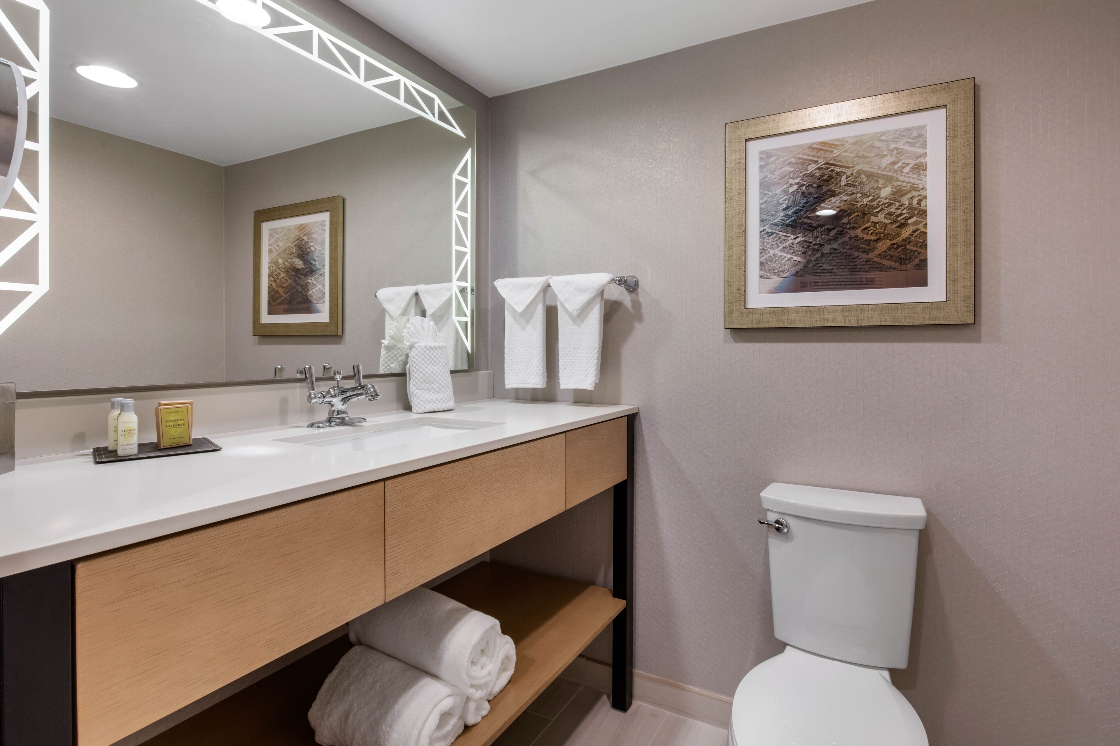 Un baño moderno con un lavamanos blanco, tocador de madera y un inodoro. Hay toallas colgadas en la pared y un cuadro enmarcado sobre el inodoro.