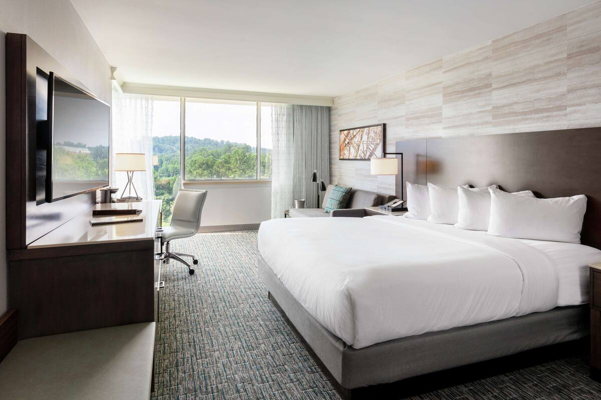 A hotel room with a large bed, a desk with a flat-screen TV, a chair, and a view of the outdoors.