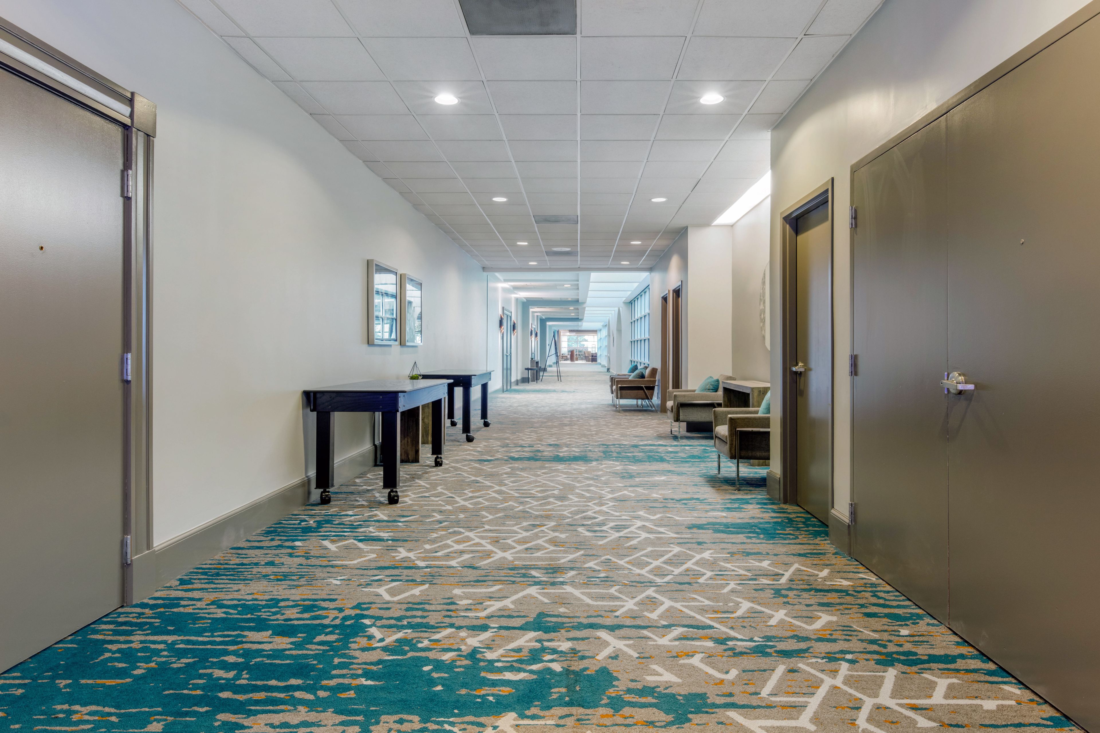 Un couloir d’hôtel avec un tapis à motifs, une longue table entourée de chaises et un coin salon avec des canapés.