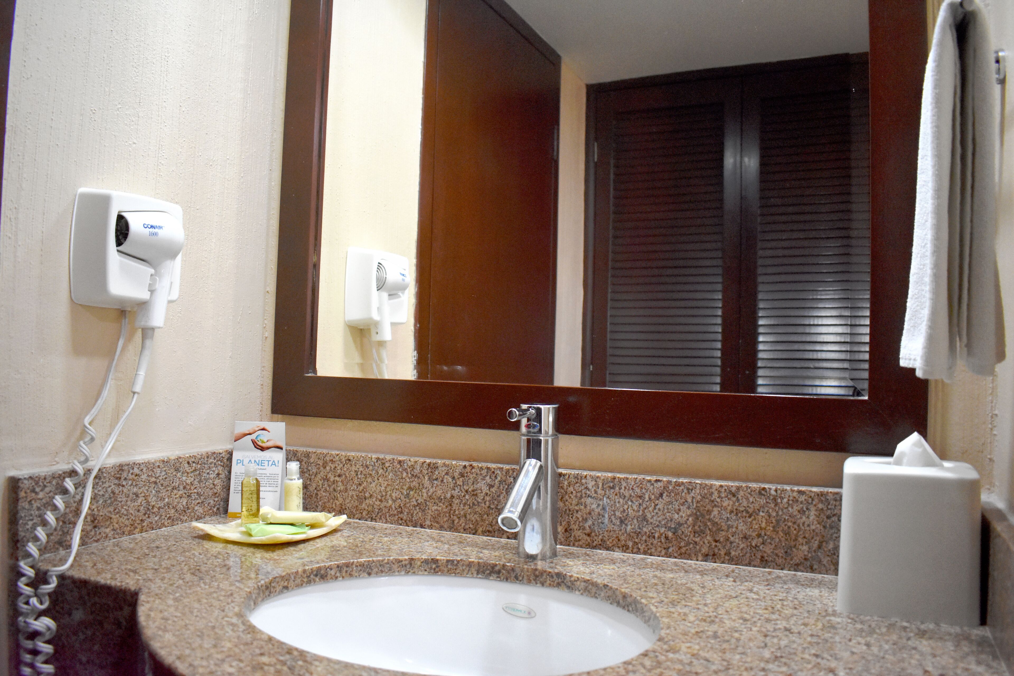 Un baño con encimera de granito, un lavabo, un espejo, un perchero para toallas y un secador de pelo fijado a la pared.