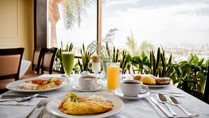 Disposição para o café da manhã com pratos de comida, café e suco, com vista para a cidade através de grandes janelas.