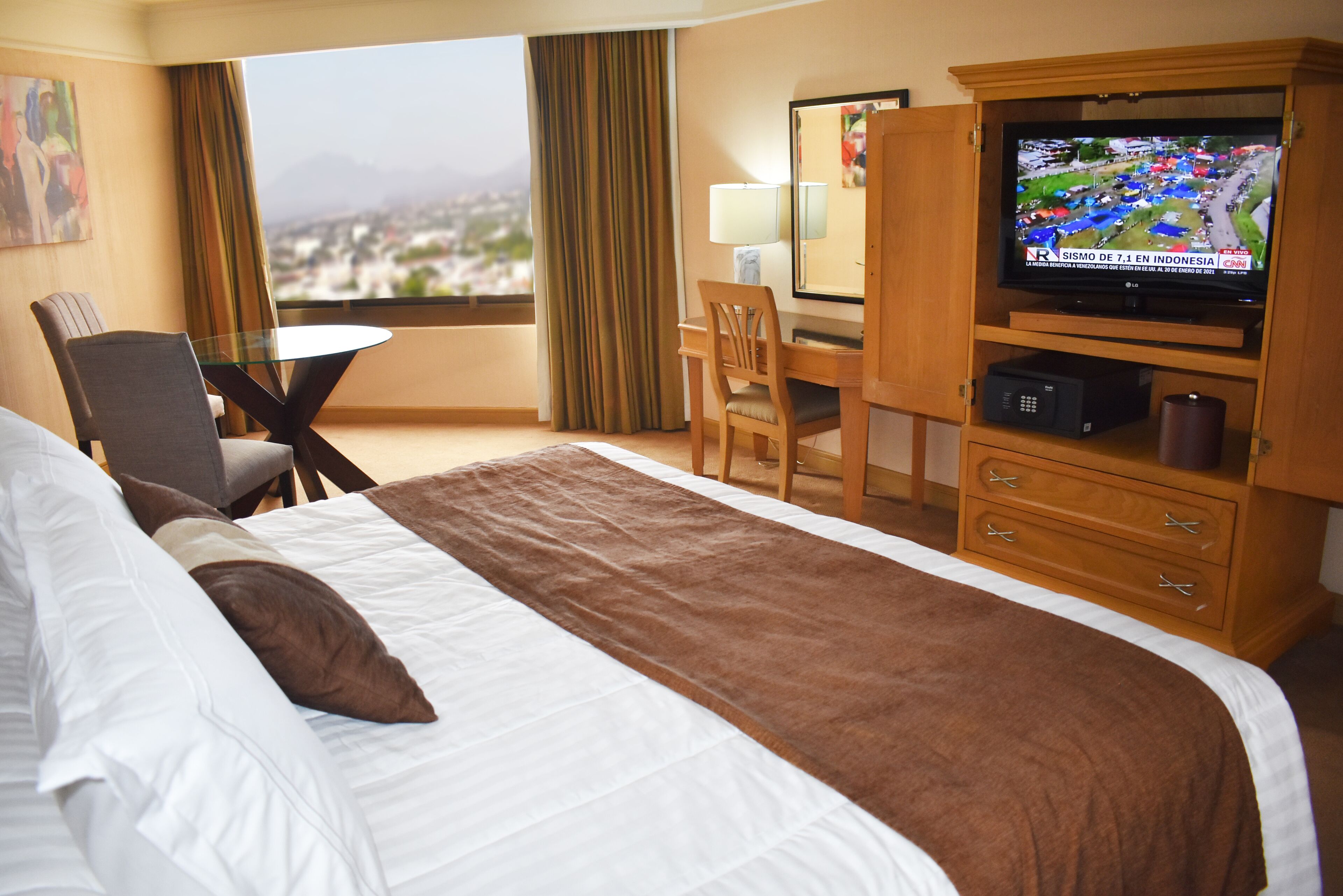 A hotel room with a large bed, a desk, a television, and a view of the city.