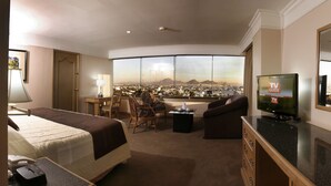 Quarto de hotel com uma cama, televisão, sofá, escrivaninha e vista para a paisagem urbana através de grandes janelas.
