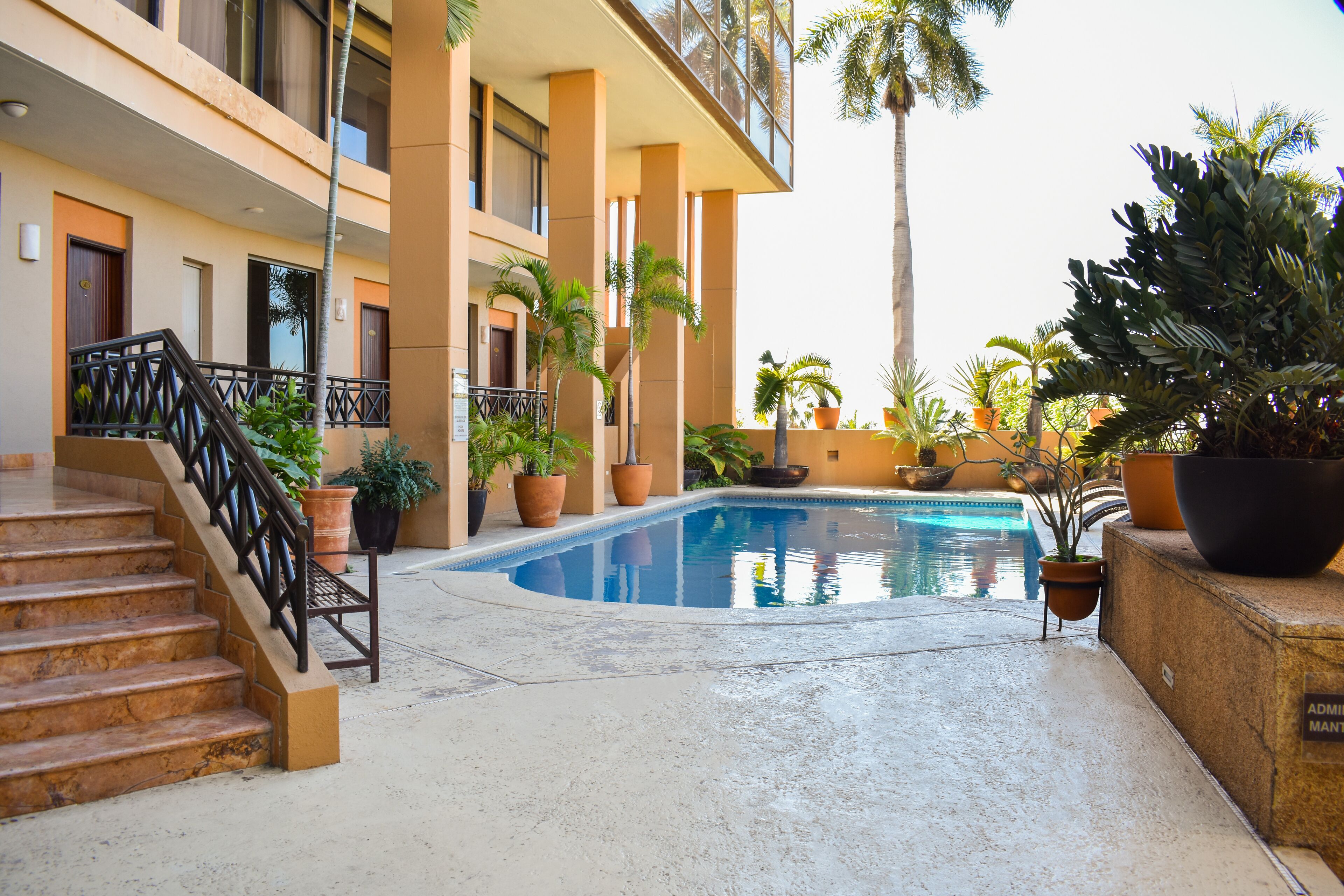 A pool area with steps leading to a balcony, surrounded by potted plants and a building with multiple floors and balconies.