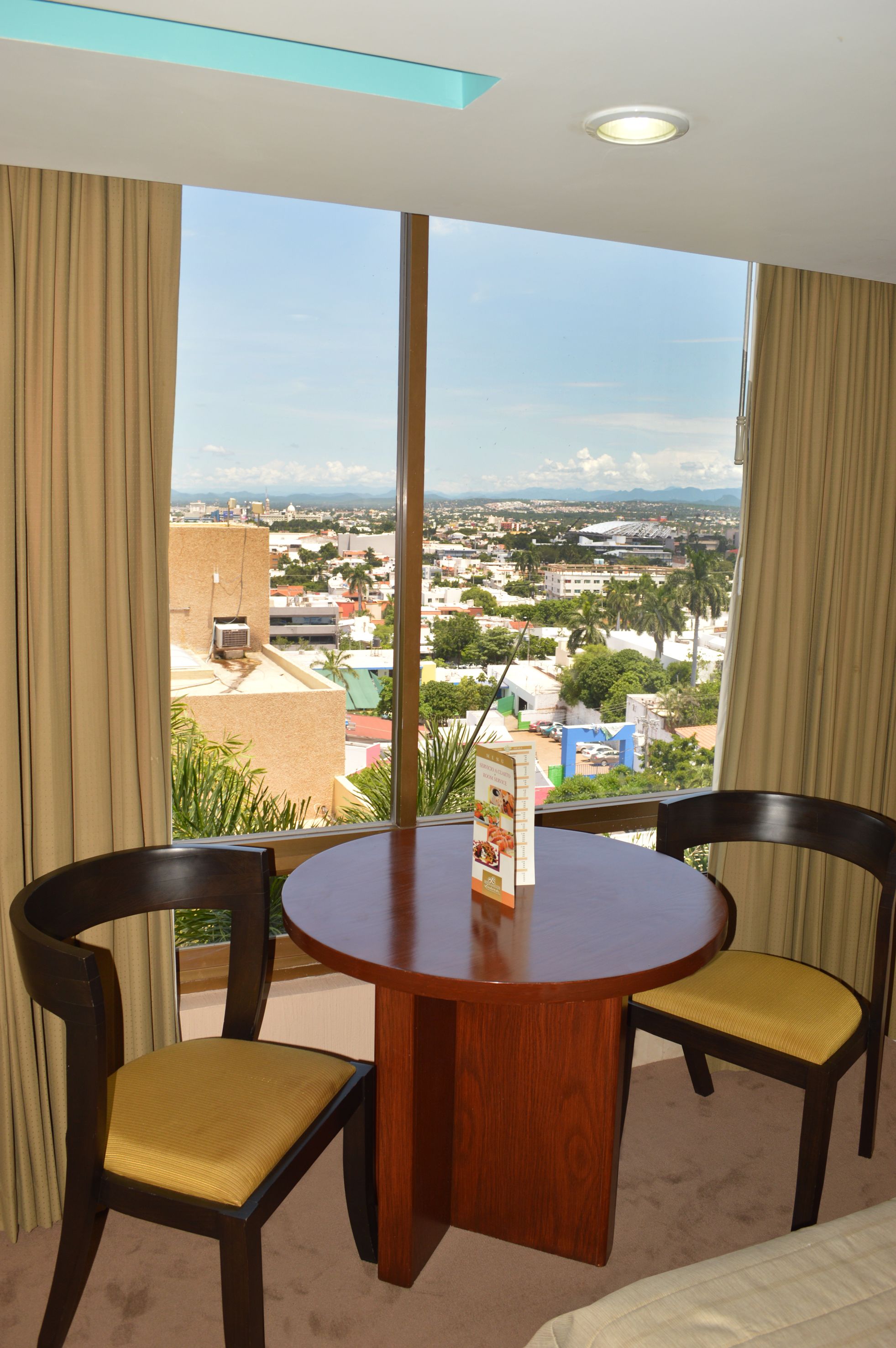 Una habitación de hotel con una mesa redonda de madera y dos sillas, un ventanal con vistas a la ciudad y cortinas color beige.