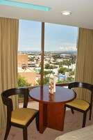Una habitación de hotel con una mesa redonda de madera y dos sillas, un ventanal con vistas a la ciudad y cortinas color beige.