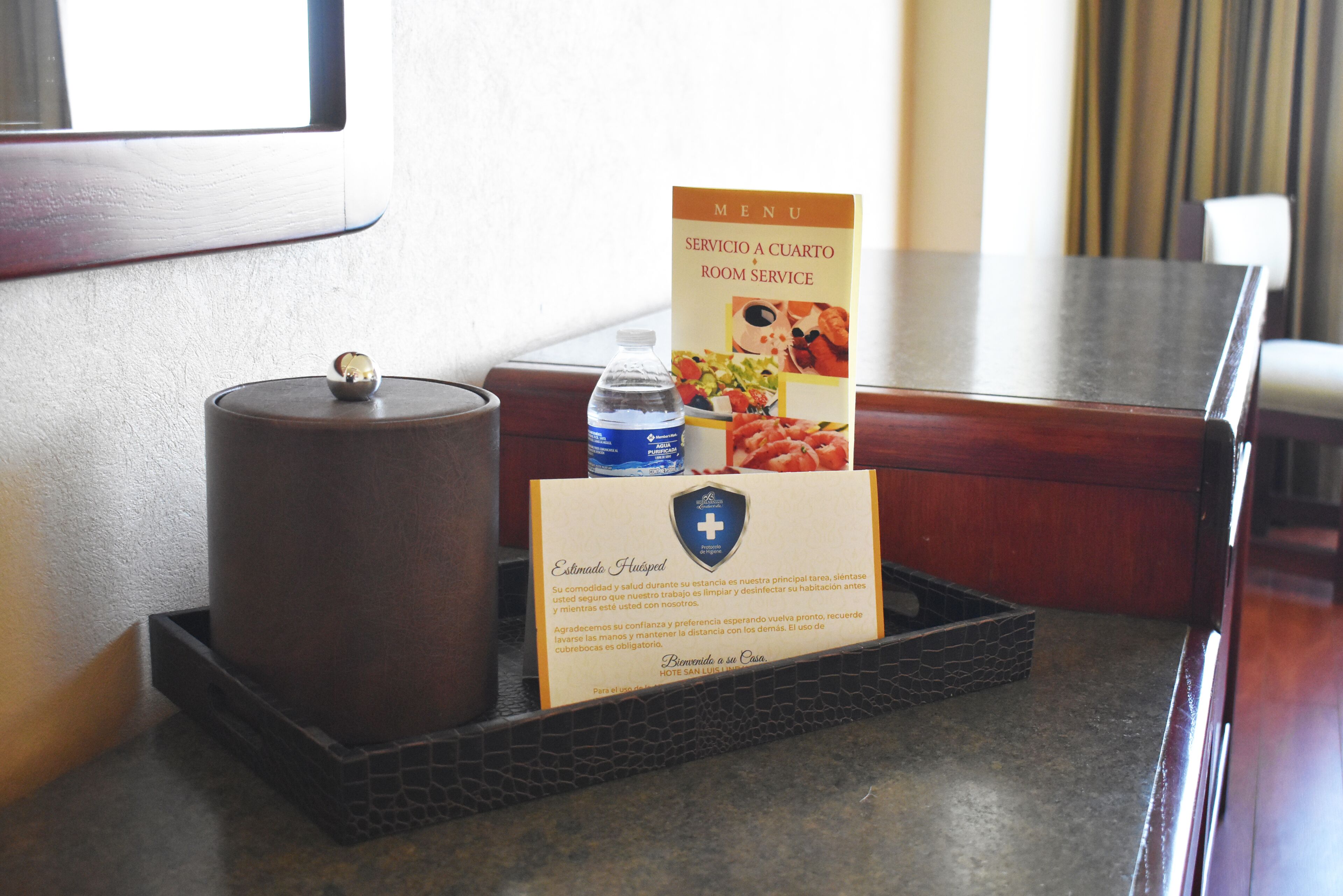 A hotel room's reception area with a wooden desk, a water bottle, a menu, and a candle.
