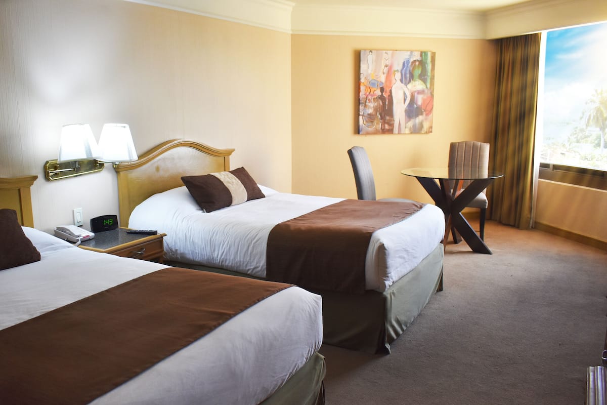 Een hotelkamer met twee bedden, een nachtkastje met een lamp, een stoel, een klein tafeltje en een schilderij aan de muur.