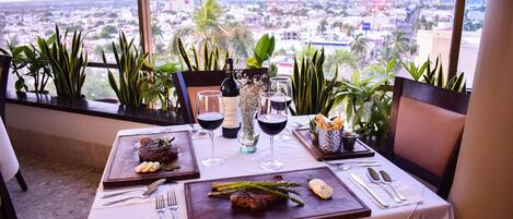 Mesa de comedor dispuesta con un bistec, espárragos y pan, con vistas a la ciudad al fondo.