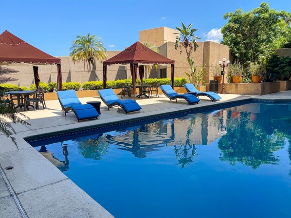 A pool area with lounge chairs and umbrellas, surrounded by greenery and a clear sky. - Hotel San Luis Lindavista (Culiacán)