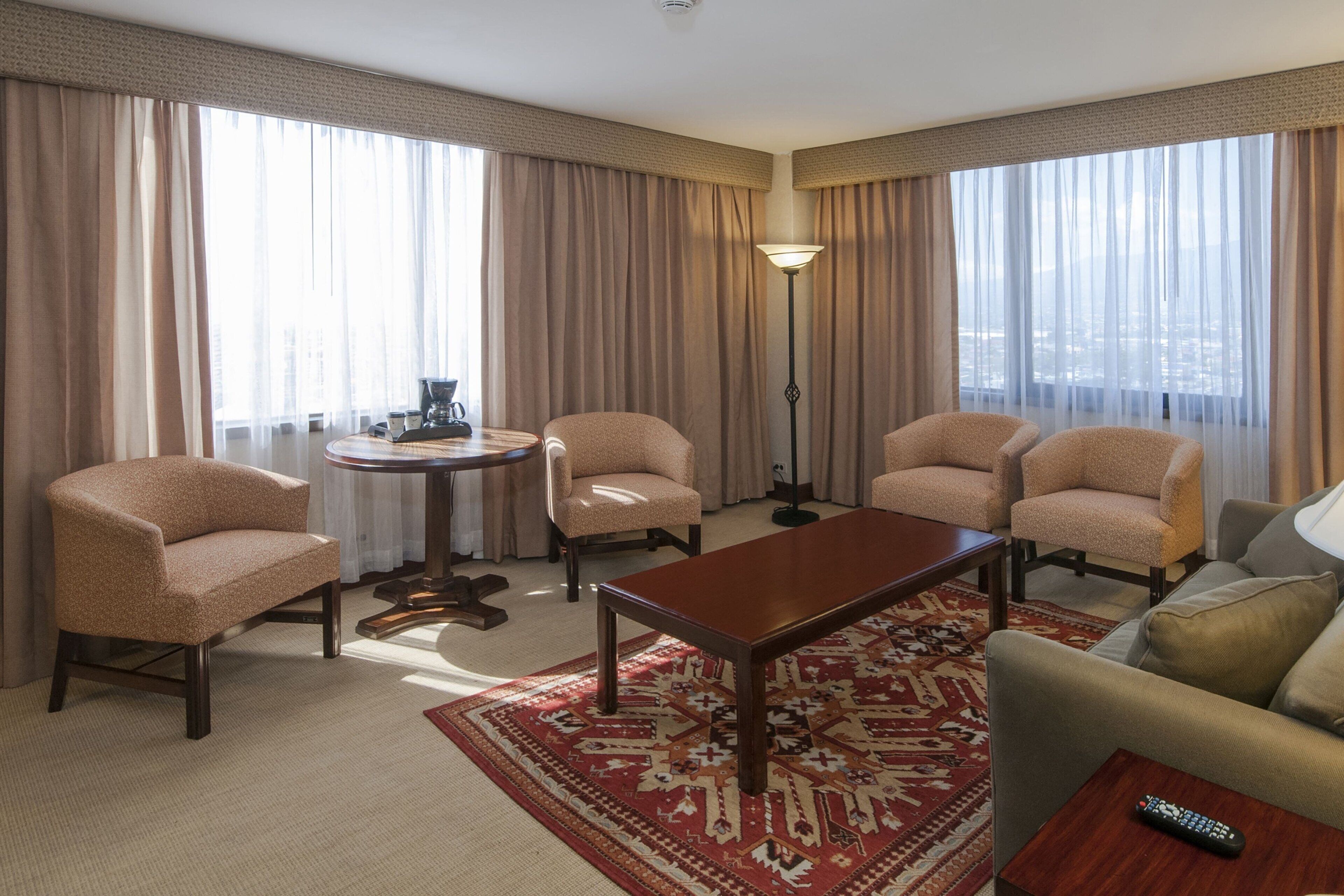 Habitación de hotel con un ventanal grande, una mesa redonda, cuatro sillones, un sofá y un control remoto sobre una mesita auxiliar.