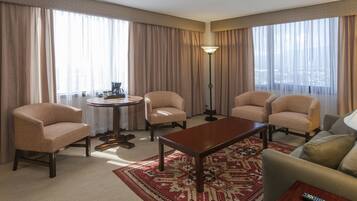 Habitación de hotel con un ventanal grande, una mesa redonda, cuatro sillones, un sofá y un control remoto sobre una mesita auxiliar.