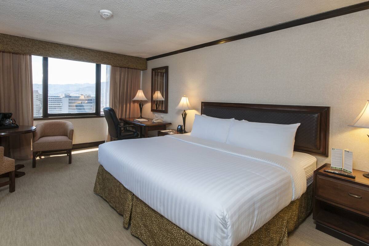 A hotel room with a large bed, a desk with a chair, a nightstand, and a window with curtains.