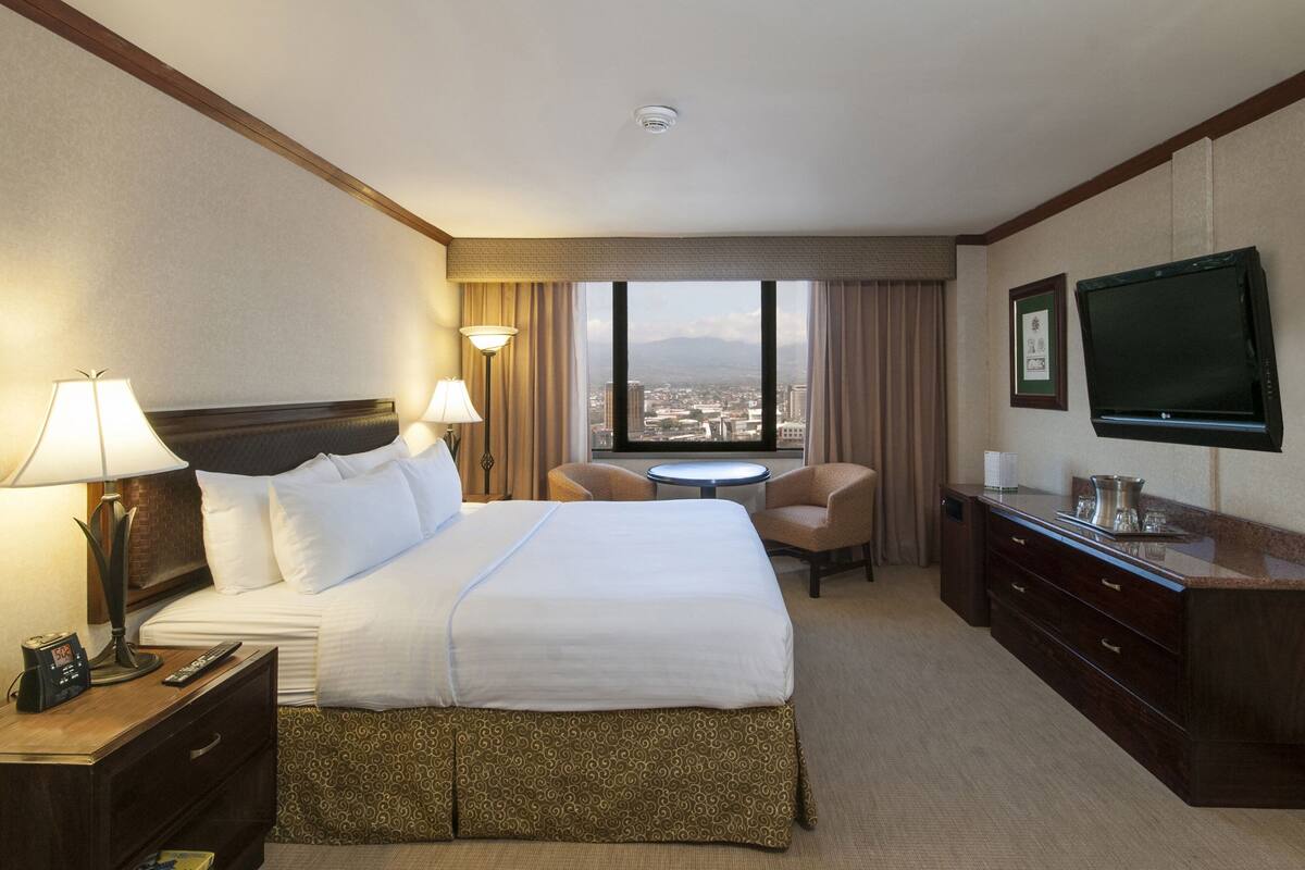 A hotel room with a large bed, two bedside lamps, a desk with a TV, a small table, and a chair.
