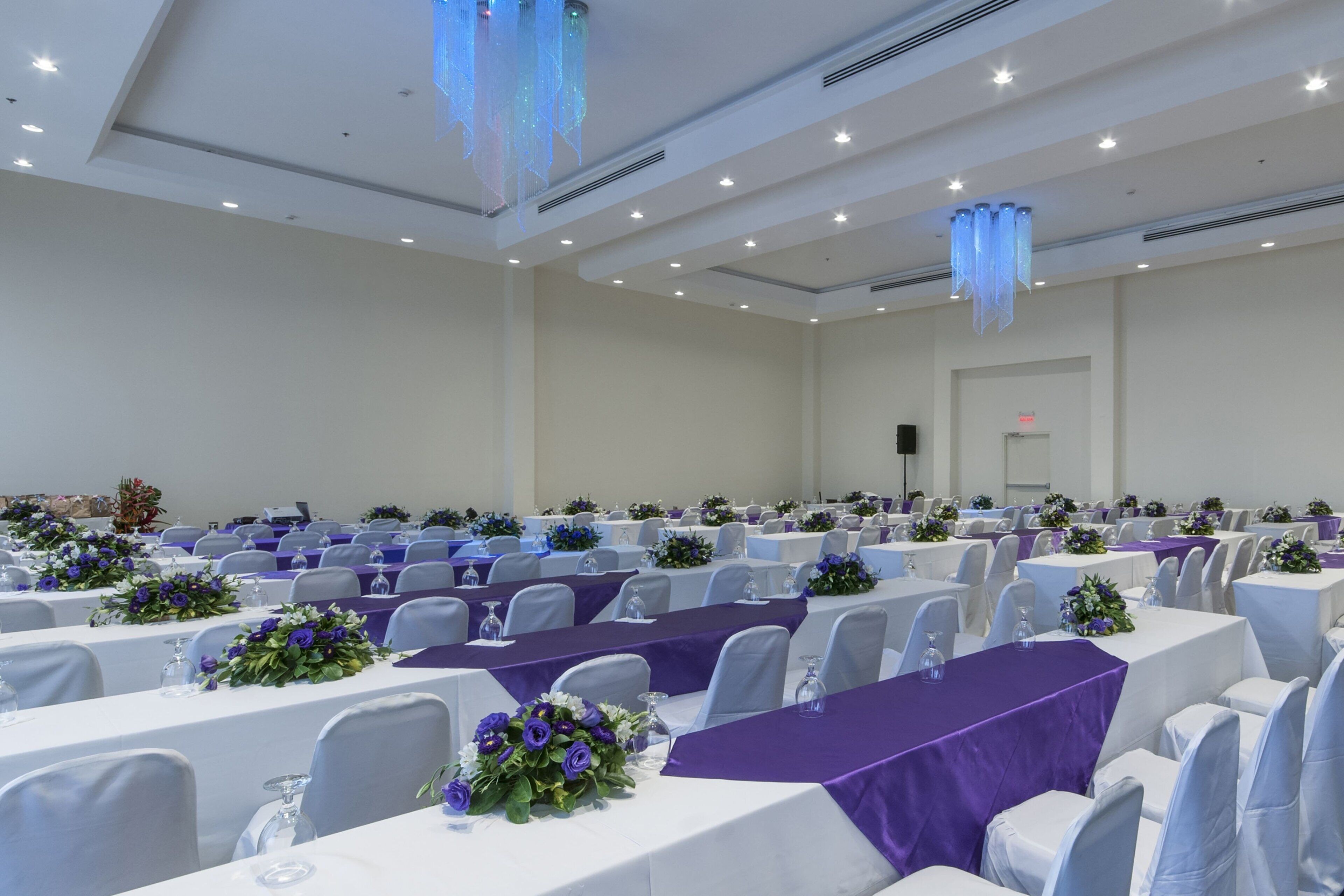 Un salón de conferencias con filas de mesas, cada una cubierta con un mantel blanco y caminos de mesa morados.