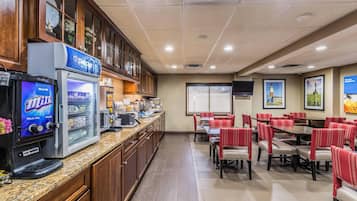 Uma área de refeições de hotel com uma máquina de café de leite integral, um refrigerador e uma tela de televisão.